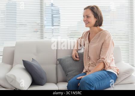 Interni luminosi con donna seduta sul divano con cuscini da persiane e luce del sole che filtra delicatamente Foto Stock