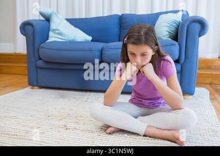 Bambina seduto a gambe incrociate su un tappeto nel soggiorno vicino al divano blu, ai cuscini e al mento riposante Foto Stock