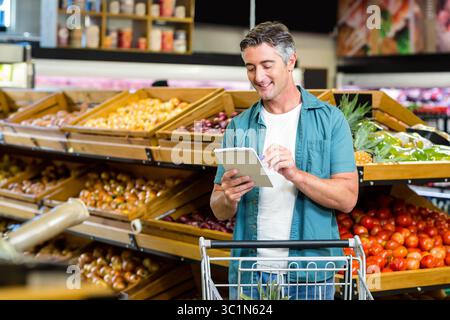 Uomo di mezza età che scrive appunti su un blocco note a spirale nel supermercato produce Aisle con carrello della spesa Foto Stock