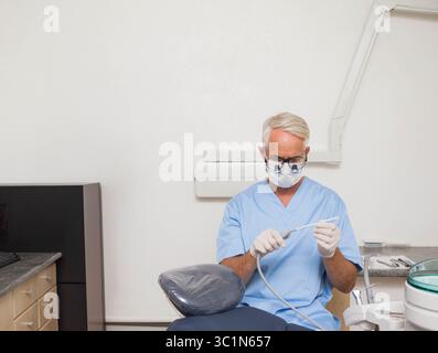 Dentista maschio anziano che indossa scrub e guanti per ispezionare il tubo di aspirazione sulla sedia dentale nella sala esame Foto Stock