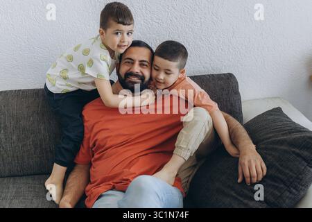 Padre felice del Medio Oriente che si rilassa sul divano con due figli che lo abbracciano. Papà sorridente che si diverte a divertirsi con i bambini a casa. Amore di famiglia, bondin Foto Stock