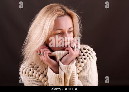 Una donna dalla pelle chiara e dai capelli biondi guarda verso il basso con un'espressione contemplativa. Indossa un maglione in maglia testurizzata color crema e io Foto Stock