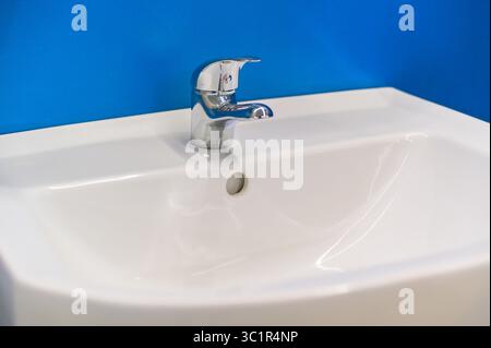 Il moderno rubinetto cromato è collocato su un lavandino in ceramica bianca in un bagno elegante con una parete blu vibrante che rappresenta l'igiene e il design degli interni Foto Stock