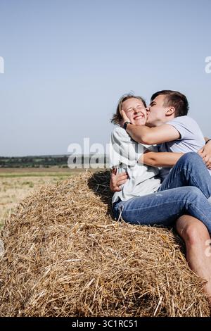Coppia romantica seduta su una balla di fieno e abbracciata in un campo rurale illuminato dal sole Foto Stock