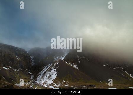 Vista suggestiva delle nuvole e della nebbia umide evidenziata dal sole che avvolge le montagne rocciose coperte di neve dell'interno dell'Islanda Foto Stock