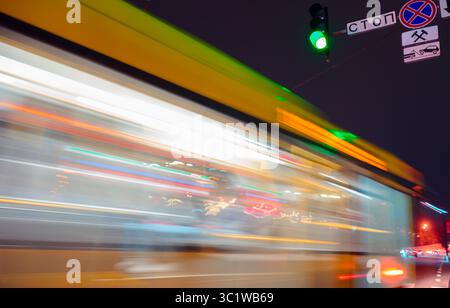 Kiev, Ucraina - 28 aprile 2025: Autobus giallo che accelera di notte sotto l'insegna del semaforo verde con effetto di sfocatura del movimento e luci colorate sullo sfondo Foto Stock