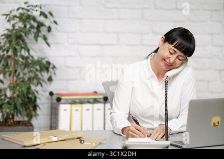 Dipendente dell'ufficio con camicetta bianca che parla al telefono e scrive su un pezzo di carta, sorridendo Foto Stock