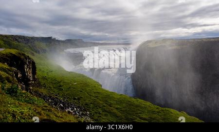 L'acqua precipita drammaticamente sul bordo della cascata Gullfoss, circondata da scogliere verdeggianti e nebbie. La luce del mattino presto crea un'atmosfera mistica, invitante esplorazione. Foto Stock
