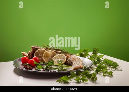 Un pesce intero splendidamente placcato poggia su un piatto nero, guarnito con erbe fresche e fette di agrumi. Il pesce è presentato su un tavolo bianco contro a. Foto Stock