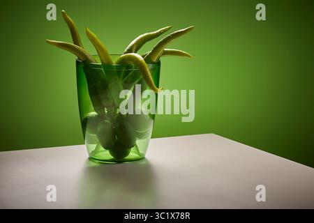Un vivace mazzo di peperoncini verdi è disposto all'interno di un vaso di vetro trasparente. Il vaso si trova su un tavolo grigio chiaro, con una parete verde fissa che funge da Foto Stock