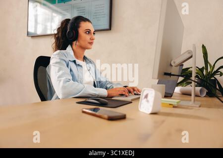 Una donna d'affari concentrata che lavora alla propria scrivania in un ambiente di ufficio moderno, che utilizza un computer desktop indossando le cuffie. Le funzioni dell'area di lavoro Foto Stock