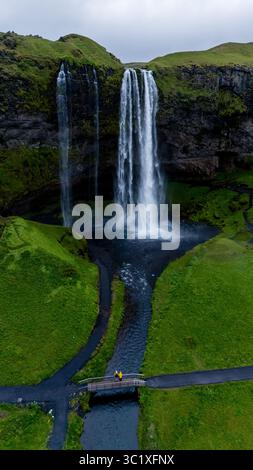 L'acqua scorre aggraziatamente dalla cascata Seljalandsfoss, circondata da vegetazione lussureggiante e scogliere rocciose. I visitatori esplorano i dintorni panoramici, apprezzando la splendida bellezza naturale dell'Islanda. Foto Stock