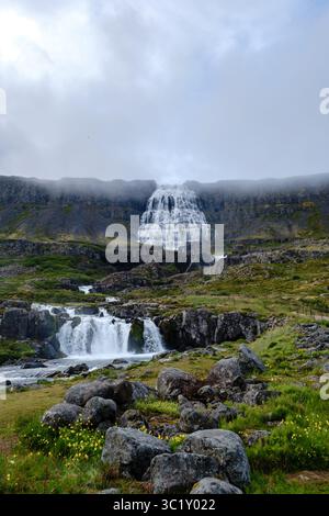 La cascata di Dynjandi, Westfjord, Islanda, crolla drammaticamente lungo le scogliere terrazzate, circondato da lussureggianti campi verdi e formazioni rocciose sotto un cielo nuvoloso in Islanda Foto Stock