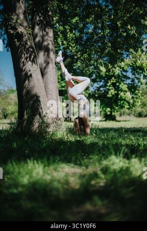 Una giovane donna che pratica fitness all'aperto e yoga facendo una testata in un parco soleggiato circondato da alberi Foto Stock