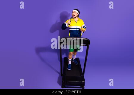 Giovane uomo che si allena su un tapis roulant mentre indossa un abbigliamento sportivo retrò, sorseggiando un drink e uno spuntino, sullo sfondo viola Foto Stock