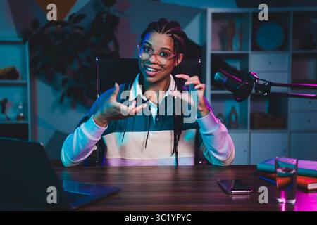 Donna affascinante che conduce una sessione podcast serale con attrezzature professionali in un moderno ufficio domestico con illuminazione vivace Foto Stock