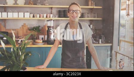 Donna in piedi con grembiule a righe che lavora dietro il bancone in legno della caffetteria, con macinacaffè Foto Stock