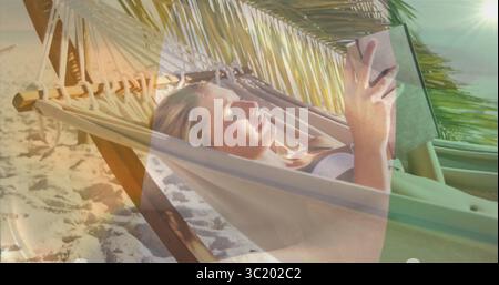 Donna che legge in costume da bagno sdraiata su un'amaca sulla spiaggia sabbiosa, tenendo un libro sotto le fronde delle palme Foto Stock