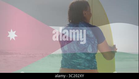 Tenendo in mano la tavola da surf gialla, donna anziana che indossa rashguard in piedi in mare con la bandiera giordana Foto Stock