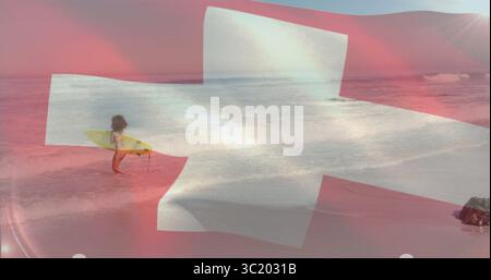 Surfista in piedi che tiene in mano una tavola da surf gialla in acque poco profonde sulla spiaggia sabbiosa, copertura della croce svizzera Foto Stock