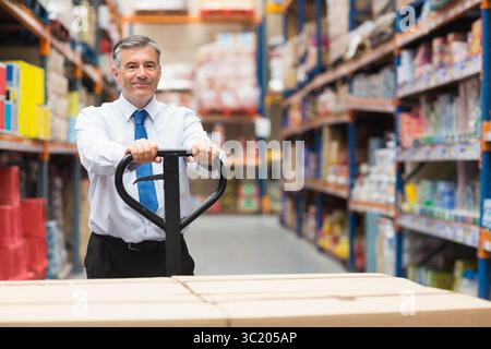 Uomo di mezza età che indossa una camicia e una cravatta che spinge le scatole per il trasporto di pallet nella corsia del magazzino Foto Stock