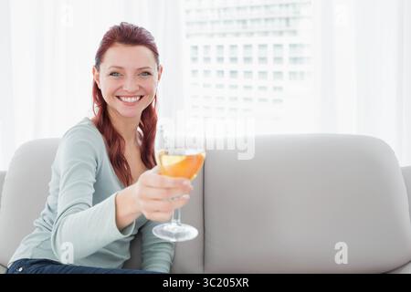 Donna dai capelli rossi seduta sul divano nel soggiorno, alzando un bicchiere di vino sulle tende delle finestre, spazio fotocopie Foto Stock