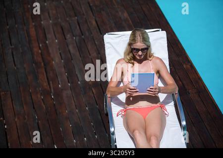 Donna sdraiata su una poltrona sul ponte della piscina, con bikini rosa e occhiali da sole in mano un tablet Foto Stock
