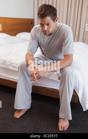 Uomo che indossa pigiami grigi seduto sul bordo del letto contro la testata di legno in camera da letto, guardando verso il basso Foto Stock