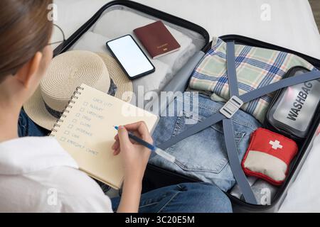 fare una lista di controllo degli oggetti da portare in viaggio. donna che scrive l'articolo della checklist sul notebook e la valigia per andare in vacanza, preparare vestiti, medicine Foto Stock