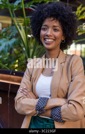 Donna afroamericana che indossa un blazer beige appoggiata su una sporgenza di legno accanto a piante in vaso nel patio del caffè Foto Stock