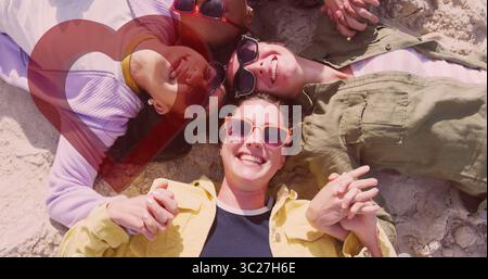 Gruppo sorridente di quattro donne sdraiate in cerchio sulla spiaggia sabbiosa, con occhiali da sole luminosi e abbigliamento estivo Foto Stock