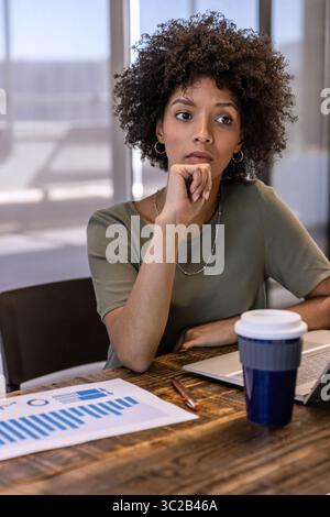Donna afroamericana che riposa il mento a portata di mano mentre studia le carte in ufficio con un portatile Foto Stock