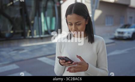 Giovane donna ispanica che utilizza lo smartphone nelle strade soleggiate all'aperto, sorridendo con fiducia mentre si impegna con la tecnologia in un ambiente urbano. Foto Stock