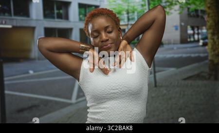 La giovane donna alza le braccia per la testa su una strada costeggiata da edifici urbani nel pomeriggio; serenità. Foto Stock