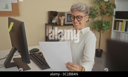 La donna tiene il documento cartaceo vicino al monitor del computer alla scrivania nell'armadio dell'ufficio; concentrazione dell'analisi. Foto Stock