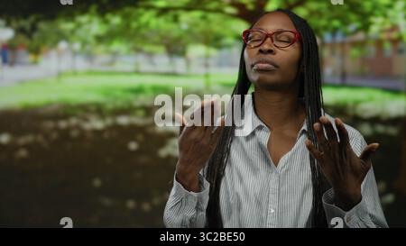 Giovane donna con capelli ricci nel parco che indossa occhiali caldi sotto il sole in un ambiente urbano all'aperto. Foto Stock