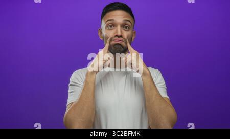 Giovane ispanico con gesti giocosi con il viso gonfiato su un vibrante sfondo viola con una camicia bianca semplice che mostra allegri espressioni Foto Stock