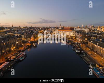 Vista aerea del tranquillo canale che riflette il soffice bagliore delle luci stradali e degli edifici, con l'iconico Royal Theatre Carré che si erge maestosamente, Amsterdam, Olanda settentrionale, Paesi Bassi. Foto Stock