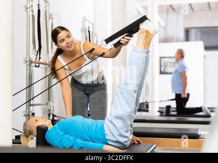 Giovane istruttrice positiva che controlla i movimenti di donna matura che esercita su attrezzature di riformatore Pilates in studio Foto Stock