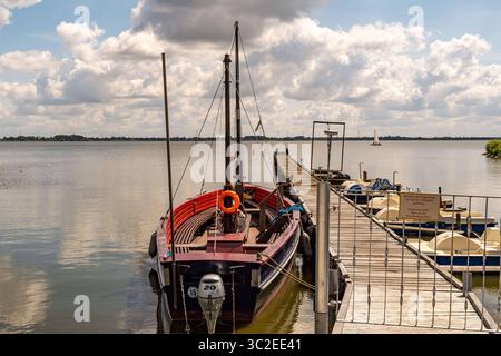 Dümmer, Deutschland 09. Juli 2025: Im Bild: Diese Nahaufnahme zeigt einen Teil eines Bootsstegs am Olgahafen des Dümmer SEE in Niedersachsen. Im Vordergrund liegt ein charakteristisches, traditionell anmutendes Boot mit Motor und Segelmast am Steg vertäut, dessen rote und dunkelblaue Elemente sich vom Wasser abheben und einen Rettungsring trägt. Daneben sind mehrere gelb-weißliche Tretboote pedalos erkennbar, die auf Freizeitvergnügen und Bootsverleih am SEE hinweisen. Der Holzsteg führt in Die weite Wasserfläche des Dümmer SEE hinaus, Die den bewölkten Himmel reflektiert. Im Hintergrund Foto Stock
