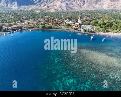 Vista aerea delle vivide acque turchesi che incontrano la riva, con barche che punteggiano l'orizzonte e montagne sullo sfondo, Pemuteran, Bali, Indonesia. Foto Stock