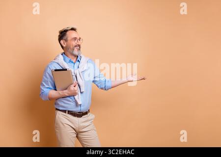 Uomo d'affari maturo in un abbigliamento casual ed elegante che presenta uno spazio per copiare, tenendo le cartelle sullo sfondo beige, con un gesto sicuro Foto Stock