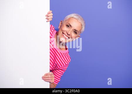 Affascinante donna matura con una camicia a righe che sbircia allegramente su una lavagna bianca su uno sfondo viola vivace Foto Stock