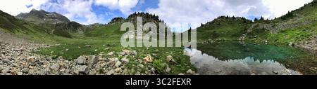 Un panorama mozzafiato che mostra un paesaggio alpino, con prati verdi, aspre cime e un tranquillo laghetto che riflette lo splendido cielo di Captu Foto Stock