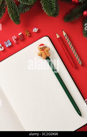 Scrivanie natalizie piatte con blocco note aperto e matite natalizie su sfondo rosso con rami di abete. Inserire il testo. Foto Stock