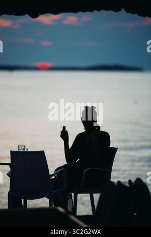 Silhouette di donna irriconoscibile seduta da sola a un tavolo in riva al mare. Bicchiere di birra sul tavolo. Sera d'estate. Fazana, Croazia - circa 2025 Foto Stock