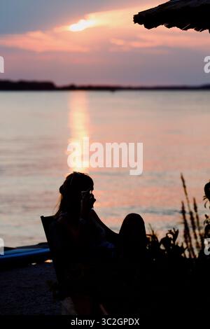 Silhouette di donna irriconoscibile seduta da sola a un tavolo in riva al mare. Bicchiere di birra sul tavolo. Sera d'estate. Fazana, Croazia - circa 2025 Foto Stock