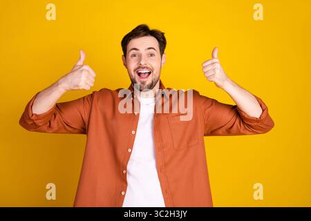 Un giovane sorridente che gestiva i pollici su sfondo giallo indossando una camicia arancione informale che esprime positività Foto Stock