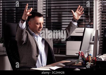 Un uomo d'affari turbato solleva le mani in frustrazione mentre lavora in un ufficio disordinato. Simbolo di esaurimento, stress sul lavoro o dimissioni. Luogo di lavoro. Foto Stock