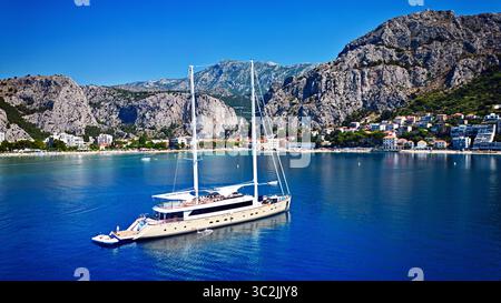 Vista aerea di un maestoso yacht che naviga sul mare turchese sullo sfondo delle aspre montagne e della città costiera di Omiš, Croazia. Foto Stock